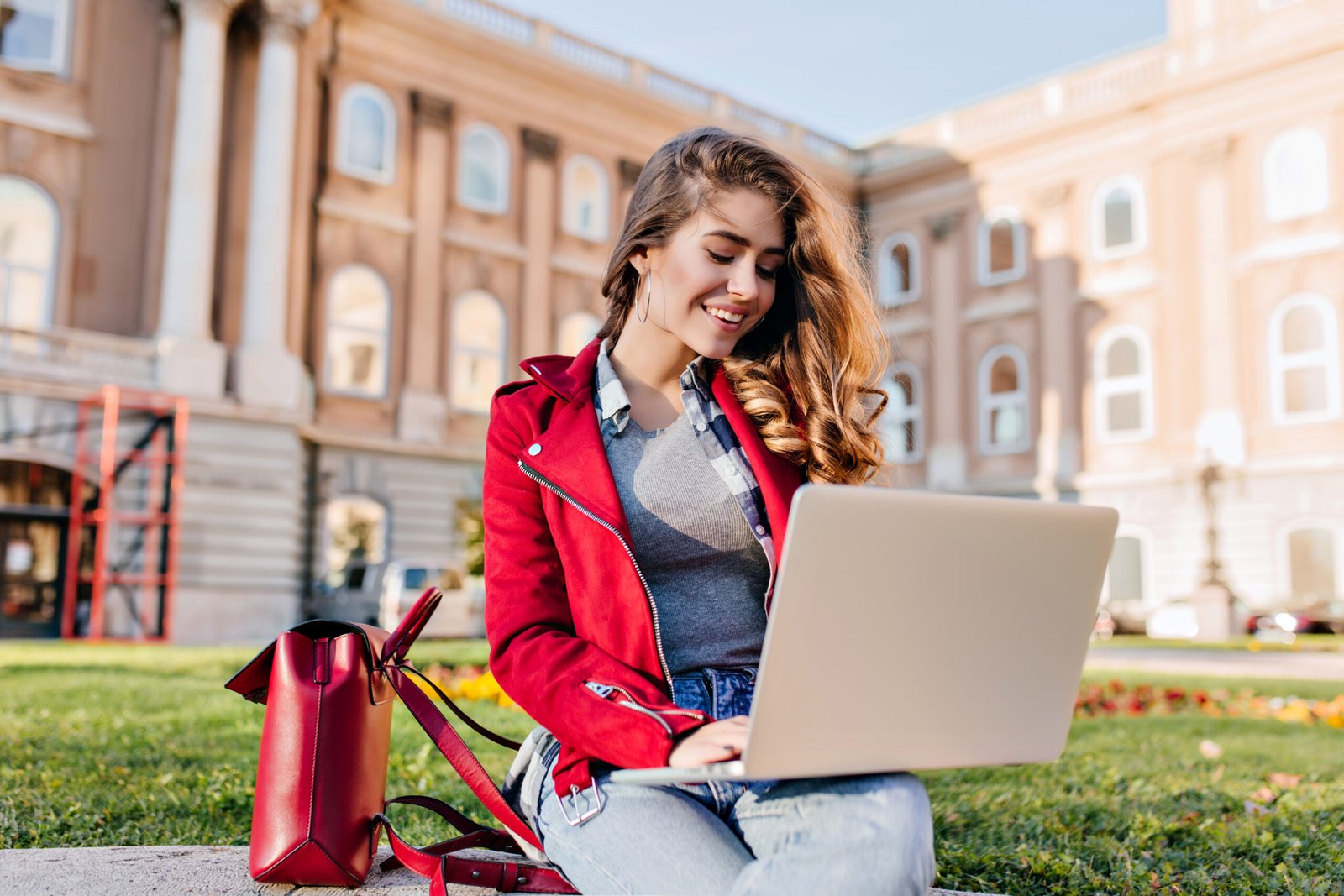 glamorous female student in red jacket sitting in the yard in front of college with computer. charming freelance worker using laptop, chilling on the grass in warm day..